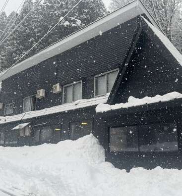 Un bâtiment moderne de couleur noire se dresse au milieu de fortes chutes de neige, entouré d'épaisses congères. Des pins s'élèvent derrière, et de doux flocons de neige tombent continuellement, créant une scène hivernale paisible.