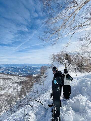 Deux snowboarders se tiennent sur la pente d'une montagne enneigée, sous un ciel d'un bleu éclatant. Des arbres dénudés encadrent la scène, tandis que des sommets lointains s'étendent à l'horizon.