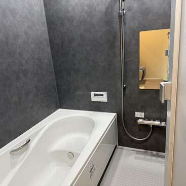 Il s'agit d'une salle de bains japonaise moderne, équipée d'une baignoire profonde, d'une douchette à main, de murs sombres, d'un miroir, d'un panneau de commande et d'un design épuré et minimaliste.