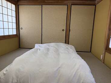 Une chambre minimaliste de style japonais, dotée de portes coulissantes traditionnelles, de tatamis et d'un lit futon avec une literie blanche, baignée d'une douce lumière naturelle filtrant à travers les cloisons shoji.