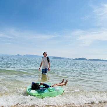 Un homme se tient dans l'eau peu profonde près du rivage tandis qu'une autre personne se détend sur un matelas gonflable vert. Ciel ensoleillé, mer calme et îles au loin.