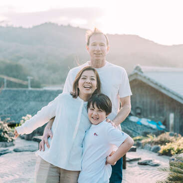Une joyeuse famille de trois personnes pose en plein air au coucher du soleil, arborant un sourire chaleureux. Ils se tiennent serrés les uns contre les autres dans un jardin paisible, avec en arrière-plan des montagnes et des maisons traditionnelles.