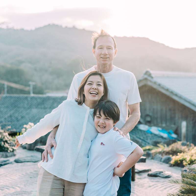 Une joyeuse famille de trois personnes pose en plein air au coucher du soleil, arborant un sourire chaleureux. Ils se tiennent serrés les uns contre les autres dans un jardin paisible, avec en arrière-plan des montagnes et des maisons traditionnelles.