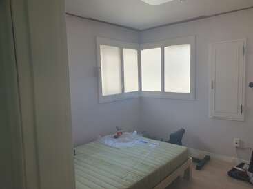 Um cômodo pequeno e claro com paredes claras tem uma janela grande, uma cama desfeita, objetos espalhados, uma cadeira e uma pequena porta na parede.