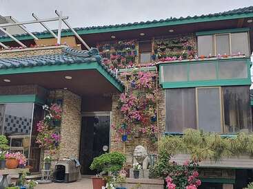 Esta imagem mostra uma casa moderna com paredes de pedra, um telhado de telhas azuis e várias flores e plantas coloridas adornando o exterior, criando uma atmosfera animada.