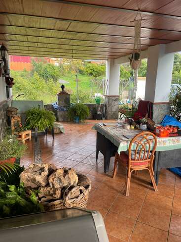 Se trata de un acogedor patio cubierto con suelo de baldosas, techo de madera, plantas, una mesa de comedor, sillas, cestas con piedras y unas vistas a un frondoso jardín.