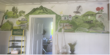 Um mural colorido retrata uma paisagem rural com montanhas, casas, árvores, animais, um avião e um pássaro, acima da porta de entrada de um cômodo aconchegante.