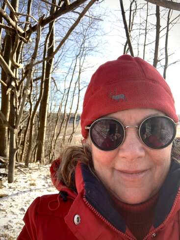 A woman in a red winter hat and coat smiles outdoors among leafless trees. She wears round sunglasses; snow lightly covers the ground under bright sunlight.