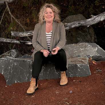 Una mujer con cabello rubio rizado está sentada en una gran piedra al aire libre, vestida con una chaqueta gris, una camisa a rayas, pantalones negros y botas marrones, sonriendo amablemente.