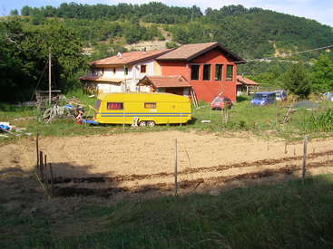 La imagen representa una escena rural con una casa roja, una caravana amarilla y un jardín en primer plano, rodeados de árboles y colinas.
