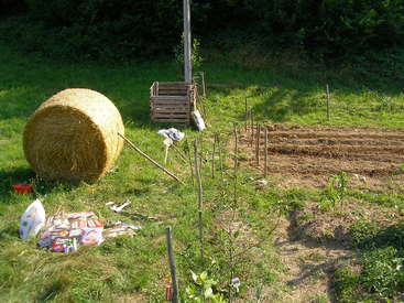 La imagen muestra un huerto con un gran fardo de heno, un cubo de abono y varias herramientas y útiles de jardinería esparcidos por el terreno.