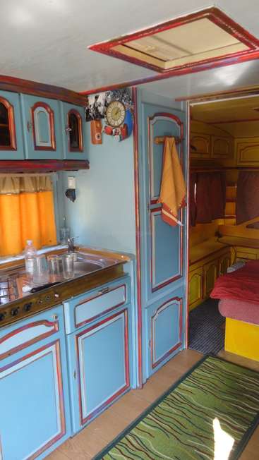 La imagen muestra el interior de una pequeña y colorida furgoneta camper, con una pequeña cocina, un fregadero y una zona de cama, con una vibrante combinación de colores y una decoración ecléctica.