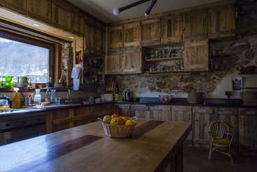 La imagen muestra una cocina con mesa y sillas de madera, una gran ventana, una chimenea y una zona de comedor, todo ello sobre un fondo de color neutro. El espacio está bien iluminado y resulta acogedor.