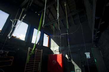 This image shows a dimly lit gym with high ceilings, ropes, a trapeze, gymnastic equipment, climbing bars, red mat, and sunlight streaming through windows.