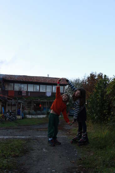 Two children stand outdoors in front of a rustic house, holding hands and forming an arch. One wears roller skates, both are smiling and playful.