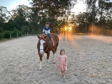 La imagen representa una serena escena de un niño montado a caballo, acompañado de una niña que lleva las riendas, sobre un fondo de árboles y una vibrante puesta de sol.
