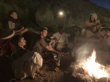 A group of people sit around a campfire at night. Some play instruments, while others listen quietly. Warm firelight illuminates their relaxed, communal gathering.