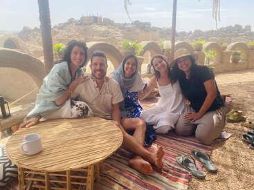 Five friends sit together on colorful rugs outdoors, smiling warmly. A round wooden table with a mug is in front of them. Desert landscape surrounds them.