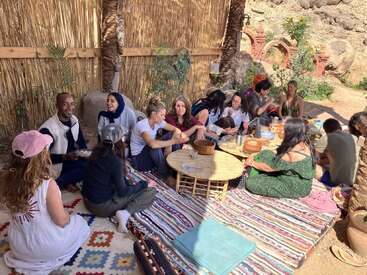 A group of people sits on colorful rugs outdoors, sharing food and conversation around low tables, surrounded by a rustic bamboo and palm fence. Relaxed atmosphere.
