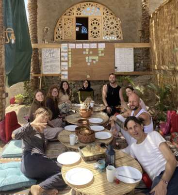 A group of people sit together around low tables outdoors, sharing food and smiling. The setting appears relaxed, cozy, and communal with decorative walls behind them.