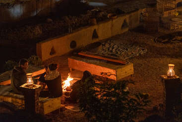 Three people sit around a small campfire at night, on wooden benches, illuminated by warm lantern light, creating a cozy and intimate outdoor setting.