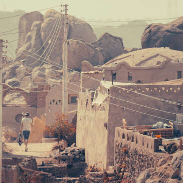 Una persona camina por un pueblo rústico con casas de adobe, rodeado de grandes formaciones rocosas y con cables eléctricos que cruzan por encima, evocando una atmósfera tranquila y antigua.