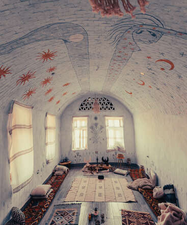 This cozy, bohemian room features floor seating, patterned rugs, soft cushions, and a high arched ceiling with celestial artwork, creating a warm, mystical, inviting atmosphere.