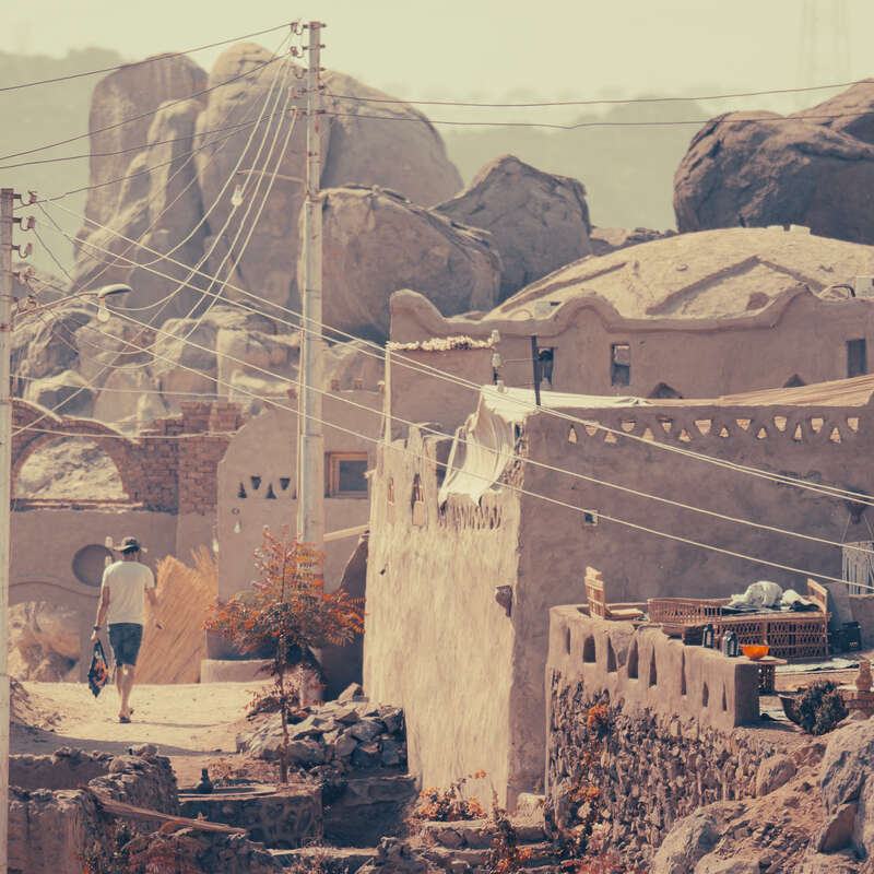 A person walks through a rustic village with adobe-style houses, surrounded by large rocky formations, power lines crossing overhead, evoking a peaceful, ancient atmosphere.