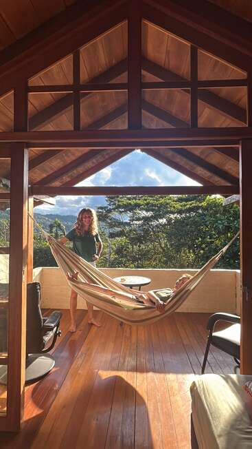 Une cabane en bois éclairée par le soleil présente un hamac confortable et deux personnes en train de se détendre, entourées d'une végétation luxuriante et d'une vue sur les montagnes, créant ainsi une atmosphère paisible et invitante.