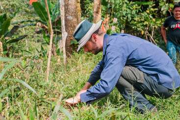 Un homme coiffé d'un chapeau et vêtu d'une chemise bleue est accroupi dans l'herbe, plantant quelque chose dans le sol. Une verdure luxuriante l'entoure ; une autre personne sourit à proximité.