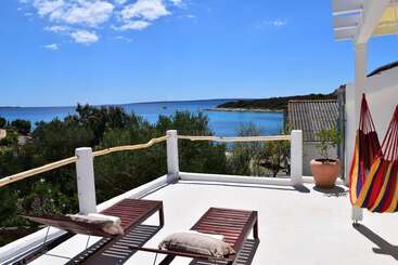 A stunning terrace overlooks a sparkling blue sea, featuring two wooden lounge chairs, colorful hammock, potted plant, sunshine, lush greenery, and relaxing, peaceful coastal scenery.