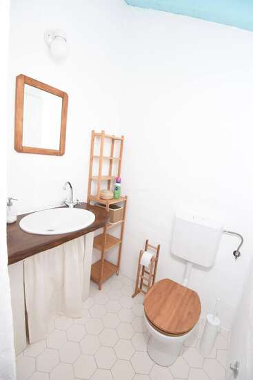 This is a small, bright bathroom with white walls, hexagonal tiles, wooden accents, toilet, sink, mirror, shelving, toilet paper holder, and minimalistic decor throughout.
