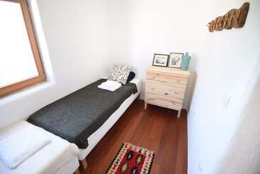 This is a small, cozy, minimalist bedroom with a single bed, wooden floors, a rug, wooden dresser, window, wall hooks, and simple decorative elements.