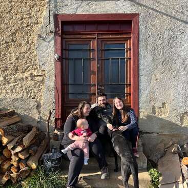 L'image représente une famille de quatre personnes, dont un bébé, assise sur les marches d'une maison rustique en pierre avec une porte et une fenêtre brunes, entourée de bois de chauffage et d'un chien.
