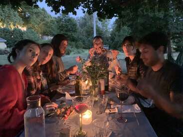 A imagem mostra um grupo de pessoas reunidas em torno de uma mesa, desfrutando de uma refeição em um ambiente externo sereno, cercado por árvores e um céu claro.
