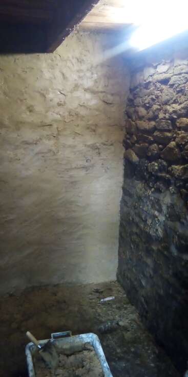 A imagem mostra um cômodo inacabado, mal iluminado, com paredes de pedra e reboco. Há materiais de construção e ferramentas no chão, o que indica um trabalho de renovação em andamento.