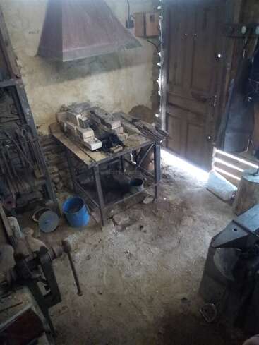 Esta é uma oficina rústica de ferreiro com uma forja, bigorna, várias ferramentas, tijolos, um torno, um balde azul e uma porta de madeira que deixa entrar a luz.