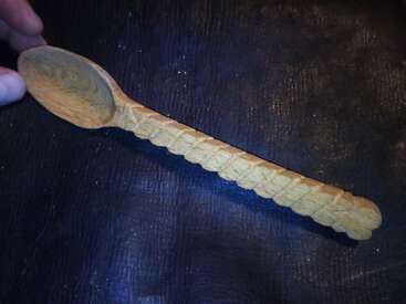 Esta imagem mostra uma colher de madeira esculpida à mão com um design exclusivo de cabo trançado. Alguém a está segurando contra um fundo de textura escura, destacando seu trabalho artesanal.