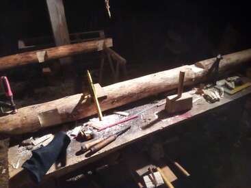 Uma bancada de marcenaria mal iluminada contém várias ferramentas, uma grande tora de madeira, grampos, formões, martelo, lápis, fita métrica, aparas e uma luva preta.