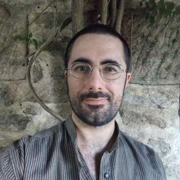 A man with glasses and a beard stands in front of a stone wall with some vines. He wears a striped shirt and smiles gently at the camera.