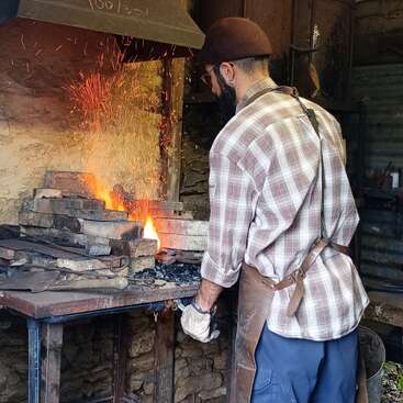 Un forgeron portant un casque et un tablier travaille dans une forge, entouré d'étincelles et de flammes, façonnant le métal dans un atelier rustique à l'ancienne.
