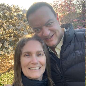 A smiling couple takes a selfie outdoors in a garden filled with blooming trees and warm sunlight, both wearing jackets and enjoying a lovely spring day.