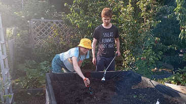 A woman in a yellow hat works in a garden bed with soil while a man in a “Spread Hummus Not Hate” shirt stands nearby watching.