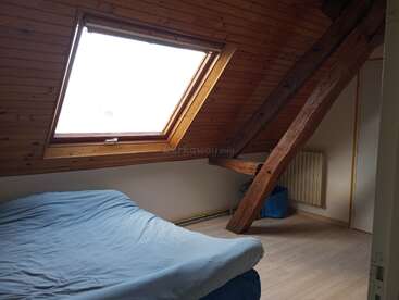 This attic bedroom features a slanted wooden ceiling, large skylight, exposed beam, simple single bed with blue bedding, wooden floor, radiator, and minimal furnishings.