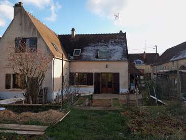 A quaint countryside house with a sloped roof, garden beds, and wooden shutters. The backyard shows traces of snow, surrounded by other cozy neighboring homes.