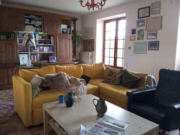 A cozy living room with a bright yellow sectional sofa, a coffee table with mugs, pictures on the wall, wooden shelves, and natural light through French doors.