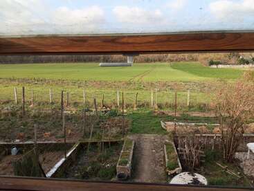 Viewed through a window, the image shows a garden with pathways, chickens in a pen, green fields, wooden fences, and a distant line of trees.