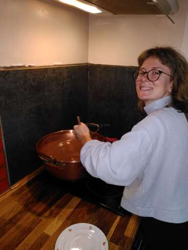 A person with glasses smiles while stirring food in a large copper pot on the stove, wearing a light sweater in a cozy kitchen setting.