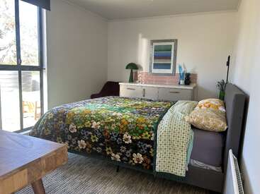 Ein gemütliches Schlafzimmer mit einem großen Fenster, einer geblümten Tagesdecke, gemusterten Kissen, einem Nachttisch, einer grünen Lampe, abstrakter Wandkunst und natürlichem Licht schafft eine friedliche Atmosphäre.