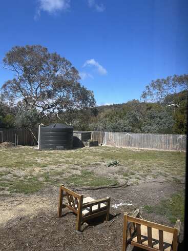 Zwei Holzstühle stehen vor einem offenen Hinterhof mit lückenhaftem Gras, einem großen Wassertank, Bäumen, einem Holzzaun, blauem Himmel und ein paar verstreuten Wolken.